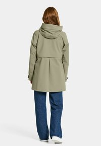 Olivi vihreä vedenpitävä takki huppulla, muotoiltu vyötärö, laskokset takana ja nepparikiinnitykset, yhdistettyinä sinisiin farkkuihin ja valkoisiin tennareihin.