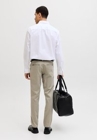 Mann i hvit skjorte og beige bukser som holder en svart duffelbag, stående foran en ensfarget hvit bakgrunn.