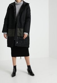 Manteau matelassé noir avec détails en panneau vert, fermeture zippée et capuche réglable ; associé à un col roulé gris et une jupe en tricot noir.
