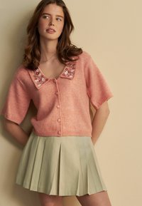 Kurzärmlige, rosa Strickjacke mit einem floral bestickten Kragen; verfügt über einen Knopfverschluss und ein kurzes Design, kombiniert mit einem plissierten, hellgrünen Rock.