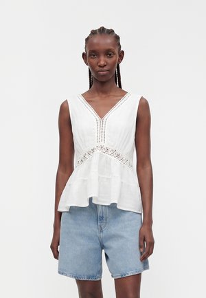 Femme aux cheveux tressés portant un haut blanc sans manches à col en V avec des détails en dentelle et un short en jean bleu clair, debout devant un fond uni.