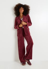 Traje de pana burdeos con una chaqueta estructurada y pantalones de pierna ancha; tela texturizada y sutiles estrías verticales. Zapatillas casuales completan el conjunto.