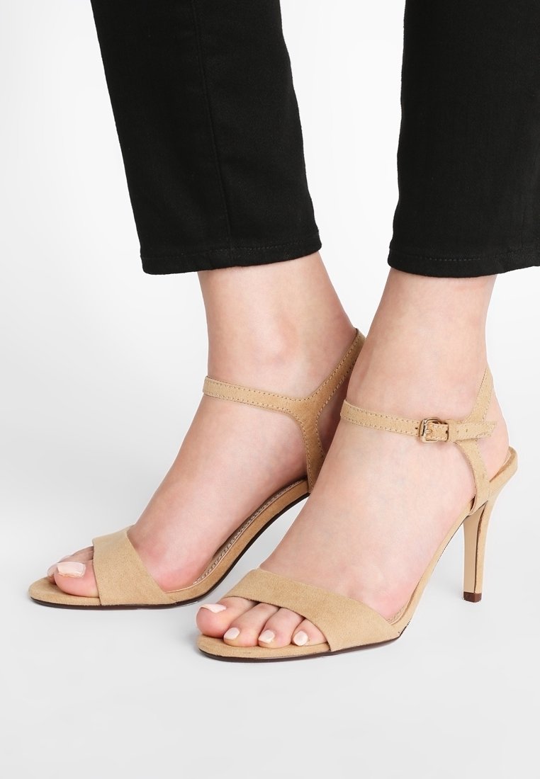 Pieds portant des escarpins en daim beige avec des brides à la cheville et un pantalon noir, posés sur un fond blanc uni.