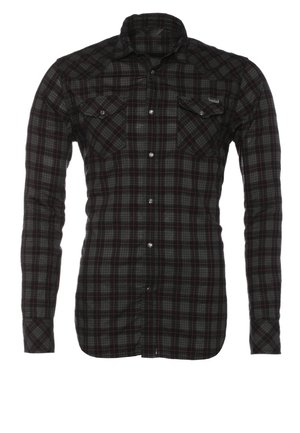 Mężczyzna idzie, ubrany w czarną koszulę w kratę na czarny t-shirt, jasnoszare dżinsy, czarne buty, nosi granatowy plecak na ramieniu.