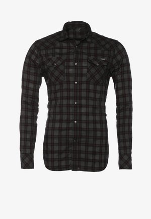 Mann geht, trägt ein schwarzes kariertes Hemd über einem schwarzen T-Shirt, hellgraue Jeans, schwarze Stiefel und trägt einen dunkelblauen Rucksack auf der Schulter.