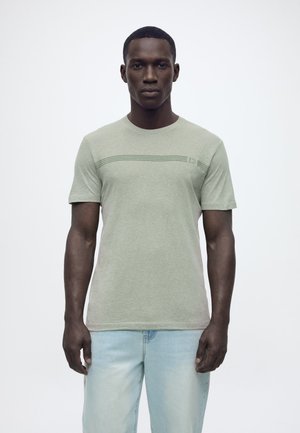 Man die een lichtgroen T-shirt met korte mouwen en subtiele horizontale strepen draagt, en lichtblauwe spijkerbroek, staand tegen een effen witte achtergrond.