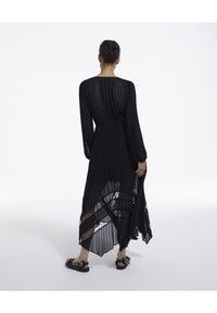Eine Frau steht mit dem Rücken zur Kamera, trägt ein langes, durchscheinendes, schwarzes Plisseekleid und schwarze flache Sandalen vor einem weißen Hintergrund.