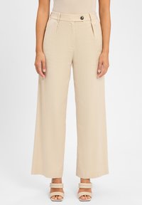 Beige wijde pijpenbroek met een gladde textuur, voorzien van een knoopsluiting en plooien aan de voorkant, gecombineerd met strappy, chunky hakken.