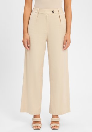 Beige wijde pijpenbroek met een gladde textuur, voorzien van een knoopsluiting en plooien aan de voorkant, gecombineerd met strappy, chunky hakken.