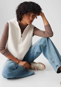 Weiße Fleece-Weste mit einem Reißverschluss vorne, über einem braunen Langarmshirt getragen. Kombiniert mit hellblauen weiten Jeans und beigen Sneakers.