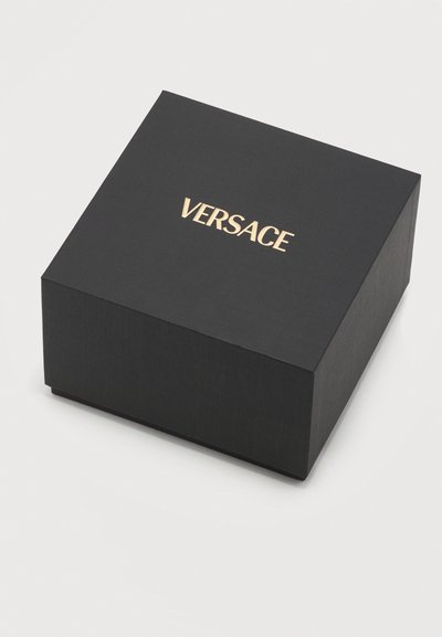 Μαύρο τετράγωνο κουτί με χρυσό λογότυπο «VERSACE» στο κέντρο του καπακιού, τοποθετημένο πάνω σε λευκή επιφάνεια.