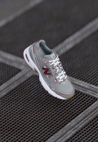 Chaussure de course pour hommes avec une tige en maille grise, des lacets blancs, des accents bordeaux et une semelle en caoutchouc avec une texture brun clair.