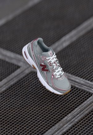 Chaussure de course pour hommes avec une tige en maille grise, des lacets blancs, des accents bordeaux et une semelle en caoutchouc avec une texture brun clair.