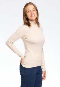 Top beige a maniche lunghe con collo alto, dalla vestibilità aderente, realizzato in materiale morbido, abbinato a jeans blu scuro con una striscia di paillettes.