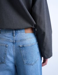 Άτομο που φοράει ανοιχτό μπλε τζιν παντελόνι με τσέπες στο πίσω μέρος και καφέ ετικέτα Topshop, συνδυασμένο με σκούρο πουκάμισο μακρυμάνικο με λεπτές κάθετες ρίγες.