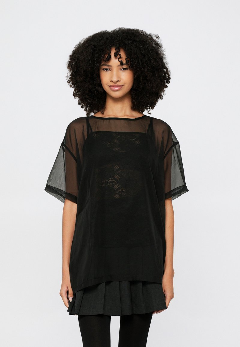 Schwarze, transparente Bluse mit kurzen Ärmeln, getragen über einem einfarbigen schwarzen Top. Verfügt über einen quadratischen Ausschnitt und eine lockere Passform. Texturiertes Design sichtbar.