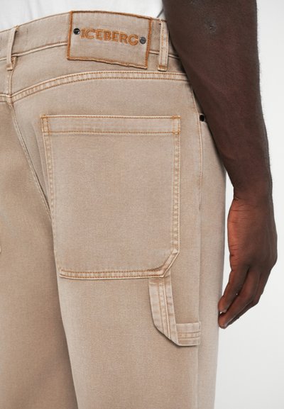 Μπεζ παντελόνι cargo με αντίθετη πορτοκαλί ραφή, διαθέτει ετικέτα στη μέση και πλαϊνή τσέπη με καπάκι.