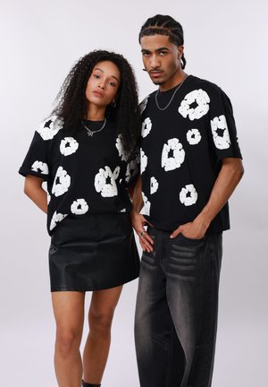 Ung kvinde og mand står side om side iført matchende sorte skjorter med store hvide abstrakte blomstermønstre.