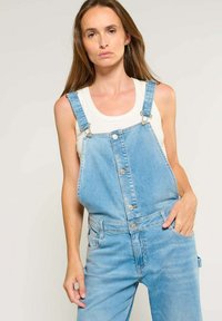 Leren denim salopette in lichtblauw met verstelbare banden, knopen aan de voorkant en zijzakken. Gemaakt om over een witte tanktop te dragen voor extra lagen.