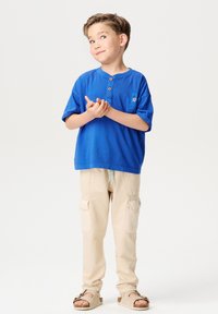 Blauwe T-shirt met korte mouwen en houten knopen, gecombineerd met beige cargobroeken met zakken. Draagt lichtbruine sandalen.