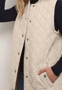 Beżowa pikowana kamizelka z zatrzaskami i bocznymi kieszeniami, z wysokim kołnierzem i teksturowanym wykończeniem. Noszona na granatowej bluzce z długim rękawem.