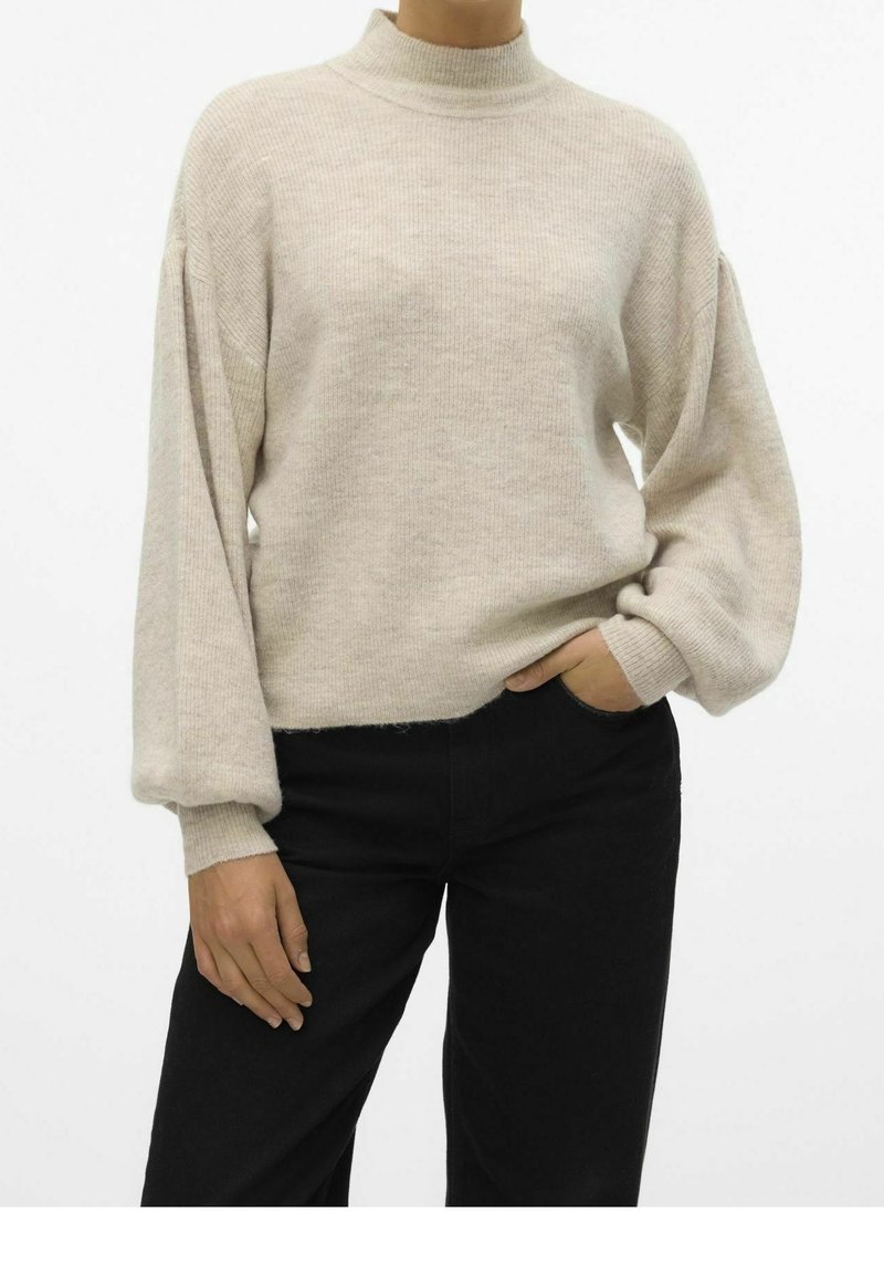 Personne portant un pull côtelé beige à manches bouffantes et un pantalon noir, une main dans la poche et l'autre détendue le long du corps.