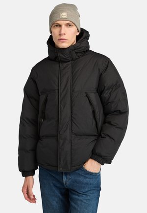 Black puffer jacket with a high collar, zippered pockets, and elastic cuffs. Topped with a light gray knit beanie.
