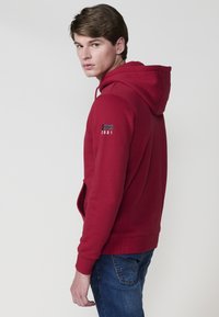 Felpa rossa con un grande tascone frontale, cappuccio regolabile con cordino e dettagli del logo sulla manica. Abbinata a jeans blu.