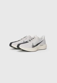 Une paire de chaussures de course Nike ZoomX Flyknit blanches et noires sur un fond blanc, inclinées vers la gauche avec des logos swoosh visibles et des semelles texturées.