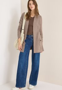 Mujer vestida con una blusa marrón, cárdigan topo, jeans de pierna ancha azules, zapatillas blancas y llevando un bolso de hombro beige, de pie contra una pared beige.