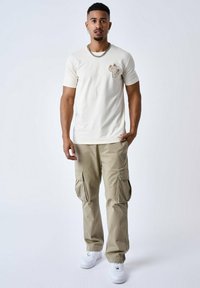 Crèmekleurig T-shirt met cactusborduursel, gecombineerd met licht khaki cargobroek met zijzakken en witte sneakers. Zilveren kettingaccessoire.