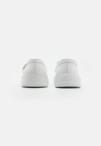 Un paio di scarpe slip-on bianche in pelle con suole spesse, viste da dietro su uno sfondo bianco semplice.