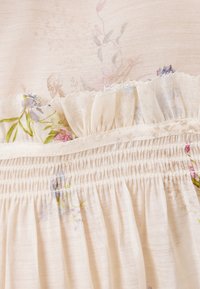 Light beige dress with floral patterns, textured ruffled trim, and smocked detailing along the waist and hem. Soft, lightweight fabric.