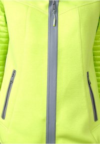 Zelená zipová bunda vyrobená z lehkého materiálu, s texturovaným bočním designem, šedými zipy a dvoma předními zipovými kapsami.