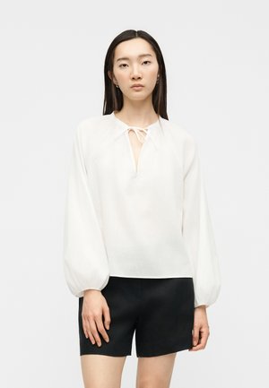 Femme portant un chemisier blanc ample à manches ballon avec un lien au col, associé à un short noir longueur genou, debout devant un fond uni.