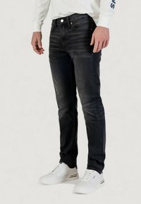 Jeans in denim nero con vestibilità slim, caratterizzati da una sfumatura sottile, design a cinque tasche, chiusura frontale con bottoni e una texture liscia. Indossati con sneakers bianche.