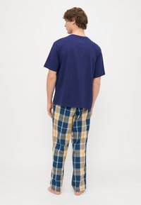 Tamsiai mėlynas medvilninis marškinėliai su apvalia iškirpte, derinami su mėlynais ir geltonais languotais kelnėmis, turinčiomis laisvą siluetą. Basas. Galinis vaizdas.