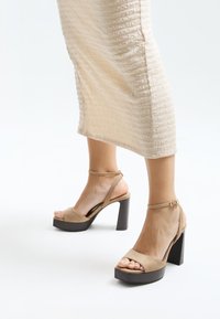 Beige suede heeled sandals with a platform sole, ankle strap, and textured brown block heel. Open-toe design with visible toes.