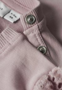 Tessuto rosa chiaro con una finitura texturizzata, caratterizzato da un bottone a pressione argento e un bordo in pizzo ondulato. Le etichette sono visibili.