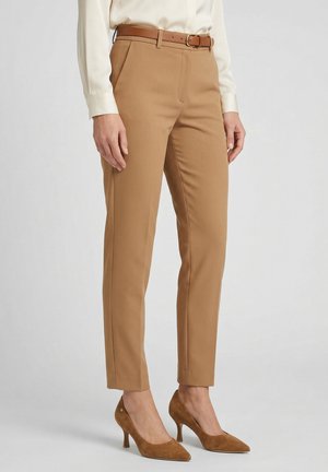 Vrouw die een slim-fit camelkleurige broek draagt met een bruine riem, gecombineerd met bijpassende bruine suède hakken en een crèmekleurige blouse.