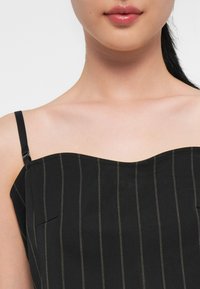 Top sin mangas negro con un escote ondulado, que presenta finas rayas verticales en dorado y un pequeño detalle de bolsillo. Tejido texturizado.