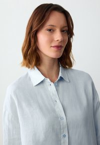 Himmelblaues Leinenhemd mit Kragen, leichter Schnitt und subtiler Textur. Kleine blaue Knöpfe entlang der Vorderblende.