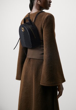 Zaino quilted nero con forma arrotondata, dettagli in metallo dorato e spallacci regolabili, indossato sopra un outfit marrone texturizzato.
