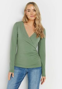 Langärmlige Wickelbluse in Hellgrün, mit V-Ausschnitt, gerafftem Seitendetail und weichem Stoff. Kombiniert mit blauen Jeans.