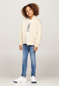 Svijetlosmeđi bomber jakna s rebrastim manžetama, nosi se preko bijele majice s grafikom. Uparen s uskim plavim trapericama i bijelim tenisicama.