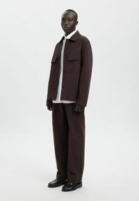 Mørk brun jakke med knappelukking og to brystlommer, kombinert med løstsittende mørkebrune bukser og svarte sko. Jevn stofftekstur.