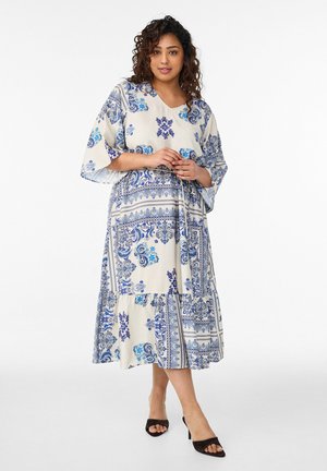 Woman standing wearing a white and blue patterned midi dress with wide sleeves and black open-toe heels against a plain white background.