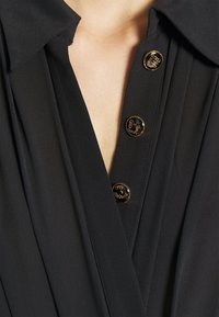 Schwarze Bluse mit klassischem Kragen, ausgestattet mit goldfarbenen Knöpfen im Kettenmuster und plissierter Textur auf dem Stoff.