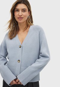 Cardigan lavorato a maglia blu chiaro con scollatura a V, tre grandi bottoni e polsini a costine. Texture morbida, vestibilità rilassata e design del collo elevato.