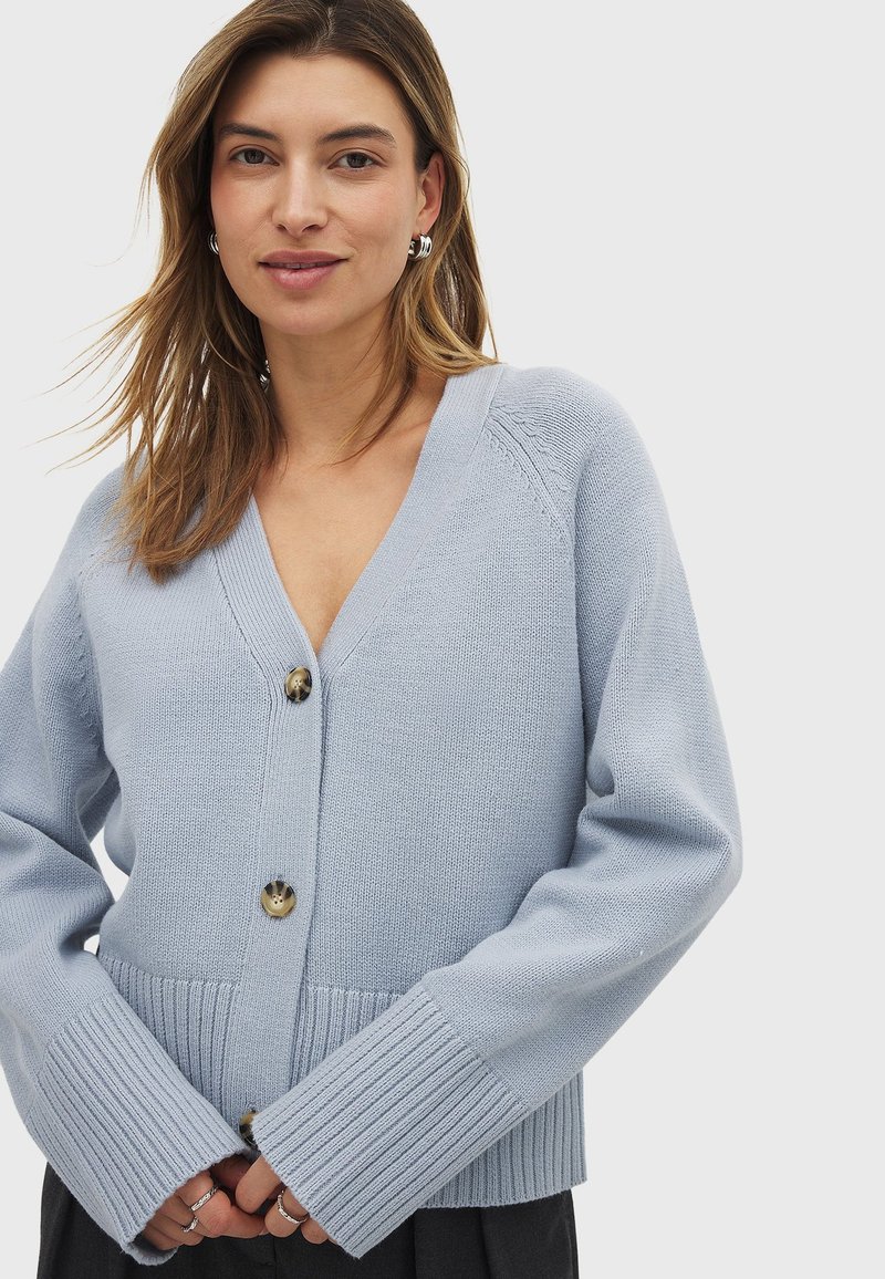 Cardigan lavorato a maglia blu chiaro con scollatura a V, tre grandi bottoni e polsini a costine. Texture morbida, vestibilità rilassata e design del collo elevato.